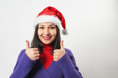 a brunette woman in a red sweater and Christmas hat gestures with her thumb up, looks at the camera and smiles. on white backgroundの写真素材