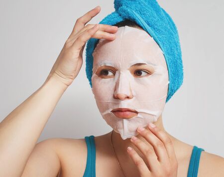 a young beautiful woman uses a moisturizing cosmetic fabric face mask with a towel wrapped around her head. on white background. cosmetic procedures, moisturizing and rejuvenation of the skin.の写真素材