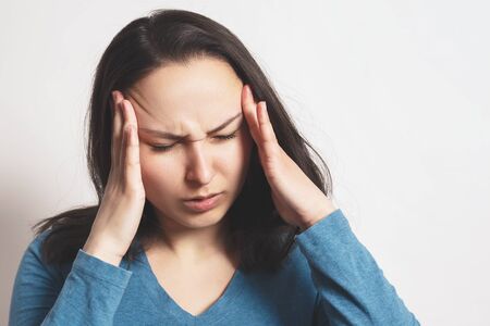 Portrait of a European young woman holding her hands to her temples with a severe headache. On light background. Migraine, high blood pressure, cardiovascular system.の写真素材