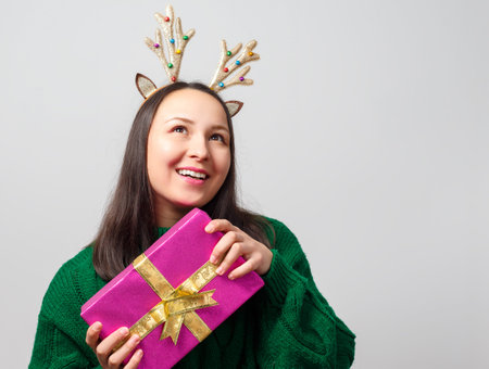 Cute young funny woman dressed as a christmas deer rejoices with a gift on a light background