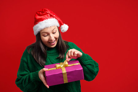 Charming woman in a Christmas cap unpacks a gift. On a red background.