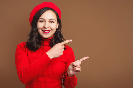 smiling young woman in red beret and sweater shows gesture on empty beige background for copy spaceの写真素材