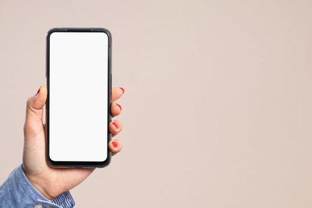 A woman's hand holds vertically a smartphone with a blank white screen on a cream background. For...