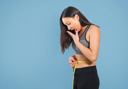 A slender woman measures her waist with a measuring tape. Against a blue background.の写真素材