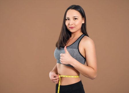 A slender woman measures her waist with a measuring tape. Against a brown background.