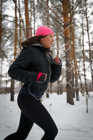Running sport woman. Female runner jogging in cold winter forest wearing warm sporty running clothing and gloves headphones. Beautiful fit female fitness model.の写真素材