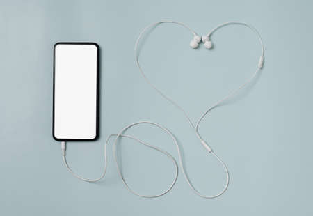 A smartphone with a white blank screen for copyspace and connected in-ear headphones in the shape of a heart, lying on a blue backgroundの写真素材