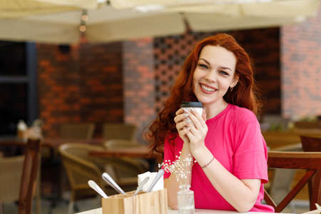 An adult red-haired woman is sitting in a summer cafe with a cup of coffee and smilingの写真素材