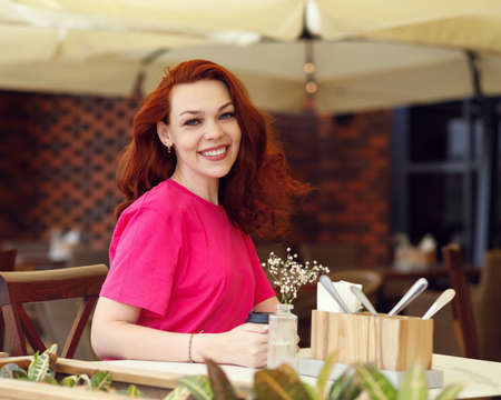 An adult red-haired woman is sitting in a summer cafe with a cup of coffee and smilingの写真素材