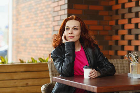 An adult red-haired woman is sitting in a summer cafe with a cup of coffee and smilingの写真素材