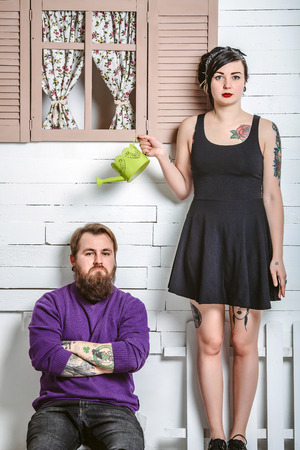 a guy and a girl with tattoos having fun, a man sits on the bench and the girl standing with a watering canの写真素材