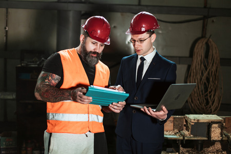Architect in suit and helmet and foreman Builder in overalls and with a tattoo compare their drawing of a construction project with a work objectの写真素材