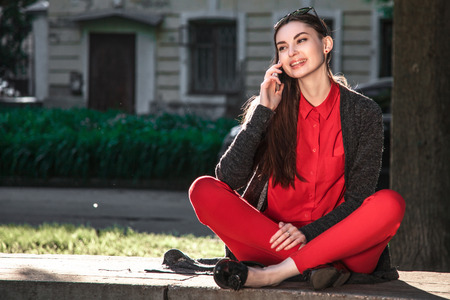 Young beautiful woman in red blouse and pants sitting in a city Park and talking on mobile phoneの写真素材