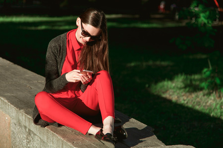 Young beautiful woman in red blouse and pants sitting in a city Park and looking to a cell phoneの写真素材