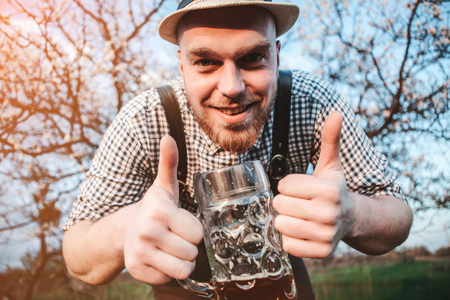 Happy smiling man with fresh brewed beer on the background of a blooming garden looking at the camera and shows that all is well. The theme is Oktoberfest, a guy in Bavarian styleの写真素材