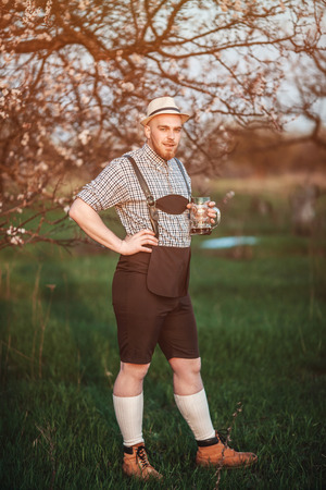 Happy smiling man tasting fresh brewed beer on the background of a blooming garden. The theme is Oktoberfest, a guy in Bavarian styleの写真素材
