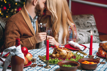 Beautiful couple in a decorated festive interior with a Christmas tree drinking wine. A romantic dinner for thanksgiving with fried chicken and candlesの写真素材