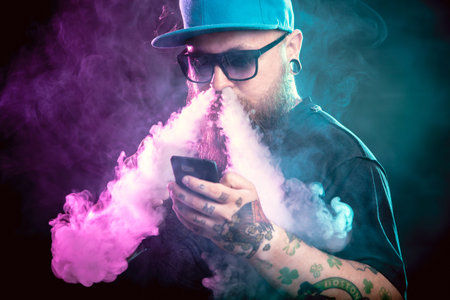 Men with beard  in sunglasses vaping and releases a cloud of vapor holding a mobile phone