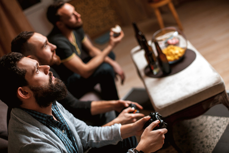 Men with a beard sitting on the couch at home with beer and chips with joysticks in hand playing computer video games. The concept of friendship, technology and weekendの写真素材