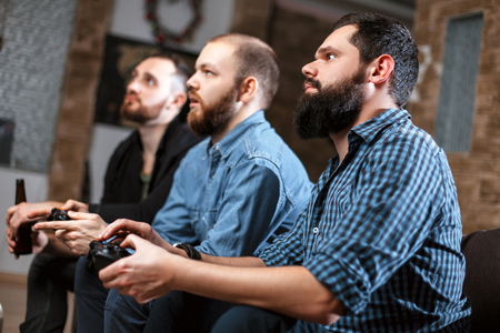 Men with a beard sitting on the couch at home with beer and chips with joysticks in hand playing computer video games. The concept of friendship, technology and weekendの写真素材