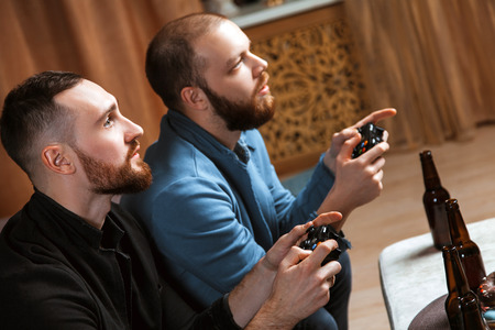 Men with a beard sitting on the couch at home with beer and chips with joysticks in hand playing computer video games. The concept of friendship, technology and weekendの写真素材