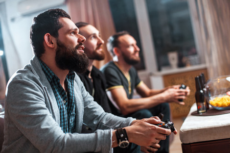 Men with a beard sitting on the couch at home with beer and chips with joysticks in hand playing computer video games. The concept of friendship, technology and weekendの写真素材