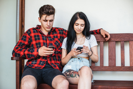 A stylish and handsome young man and girl sitting on a bench and staring at their mobile phones. Concept internet, social media network, virtual communicationの写真素材