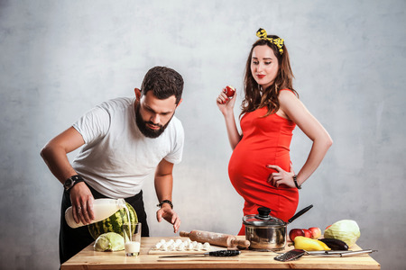 A beautiful pregnant woman with big belly in a red dress is standing, eating an apple and looking at her husband who is cooking for her beloved wife vareniki. Couple in love, family life, values, character, leaderの写真素材