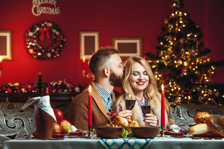 Beautiful couple kissing and holding glass of wine in a decorated festive interior with a Christmas tree. A romantic dinner for thanksgiving with fried chicken and candlesの写真素材