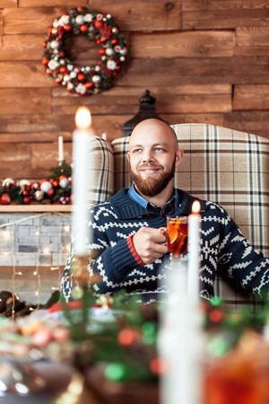 A man with a beard sitting in a chair with a Cup of mulled wine at the dinner table on Christmas. The concept of a family holidayの写真素材