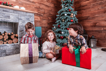 Beautiful children playing with gifts in hand in a Christmas interior with Christmas tree and fireplace. The concept of a family holidayの写真素材