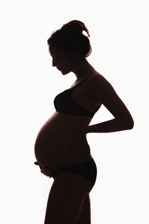 A beautiful pregnant woman with a big belly with her husband husband sitting on the couch and watching TV, a remote control in the hands of a man. Couple in love, family life, values, children, spousesの写真素材