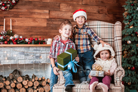 Beautiful children playing with gifts in hand on the armchair in a Christmas interior with Christmas tree and fireplace. The concept of a family holidayの写真素材