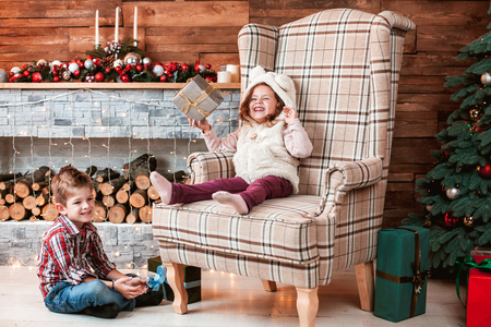 Beautiful children playing with gifts in hand on the armchair in a Christmas interior with Christmas tree and fireplace. The concept of a family holidayの写真素材