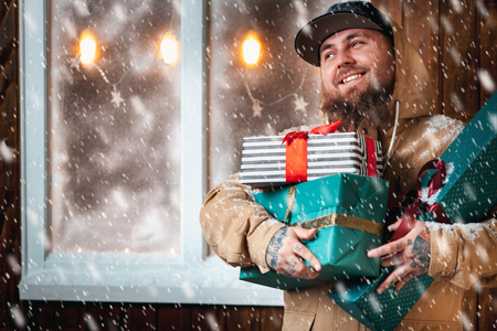 Christmas night, snow falling, funny man with a beard and with gifts in hands by the window. Winter background with lit garlandsの写真素材