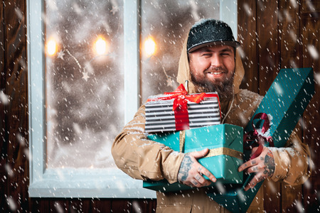 Christmas night, snow falling, funny man with a beard and with gifts in hands by the window. Winter background with lit garlandsの写真素材