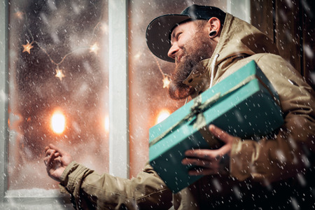 Christmas night, snow falling, funny man with a beard and with gifts in hands by the window. Winter background with lit garlandsの写真素材