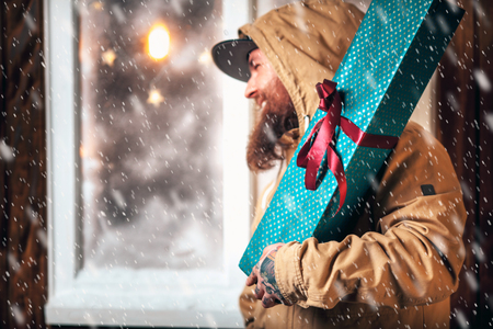 Christmas night, snow falling, funny man with a beard and with gifts in hands by the window. Winter background with lit garlandsの写真素材