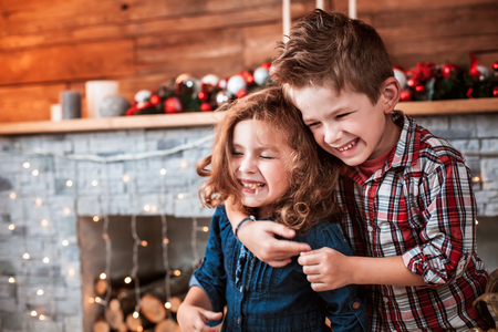 Beautiful happy laughing children in New Year interior with Christmas tree and fireplace. Brother hugging his sister. The concept of a family holidayの写真素材