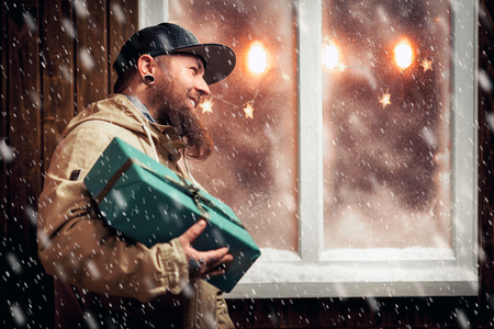 Christmas night, snow falling, funny man with a beard and with gifts in hands by the window. Winter background with lit garlandsの写真素材