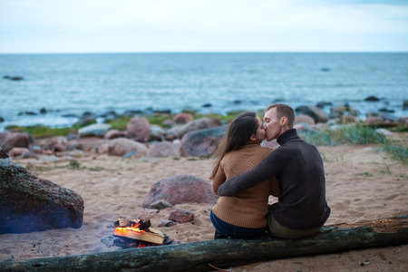 Loving couple sitting back on a log by the fire on the rocky beachの写真素材