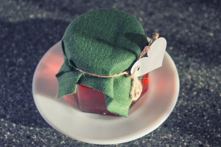 Glass jar with honey covered with green cloth with thread with white paper heart for Valentine's Day. White plate.の写真素材