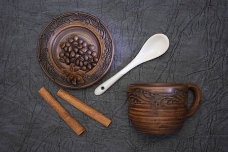 Home-made cup and saucer of burnt glazed clay. Brown ceramics on the background of a cut of dark gray dermantine. Cinnamon sticks. A pinch of brown roasted aromatic coffee beans. White glazed porcelain coffee spoon.の写真素材