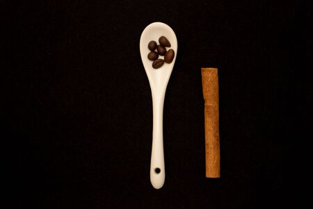 White glazed porcelain coffee spoon on a dark velvet background. Spoon with a hole in the handle. Roasted coffee beans in a spoon. Cinnamon sticks.の写真素材