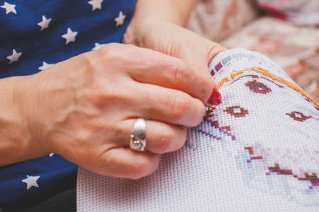Favorite activity during the pandemic. A woman sitting on a sofa is embroidering with a cross on a canvas with a pattern. Woman's hands.の写真素材