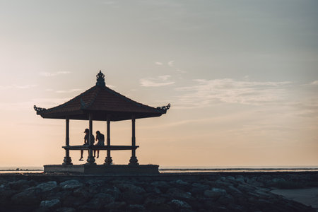 Silhouette of a relaxed couple in a gazebo on the island of Bali watching a beautiful sunsetの写真素材