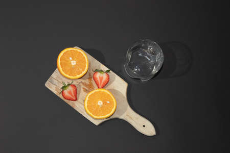 Summer scene with orange and strawberry on a wooden board with glass of water, on a black background. Minimal aesthetic.の写真素材