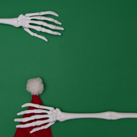 The skeleton's hand holds Santa's hat. Green background with copy space. Minimal flat lay concept.