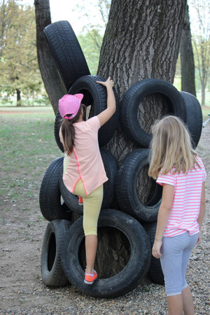 Two curious toddlers in their play clothes and bare feet joyfully scale a tire-laced tree in the park, their laughter ringing out across the playground and grassy ground belowの写真素材