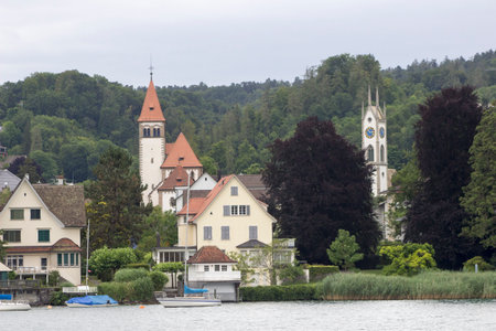 A tranquil town basks in the warmth of the sun, with a sky full of fluffy clouds and trees framing an array of buildings alongside a river or lake, with a few boats or watercrafts peacefully drifting along its waterwayの写真素材